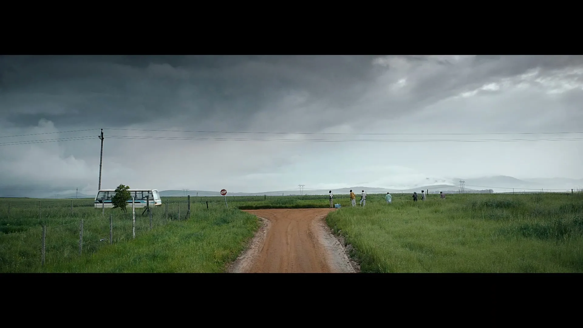 A group of people walk along a dirt road in a vast green field under a cloudy sky, with a bus parked nearby.