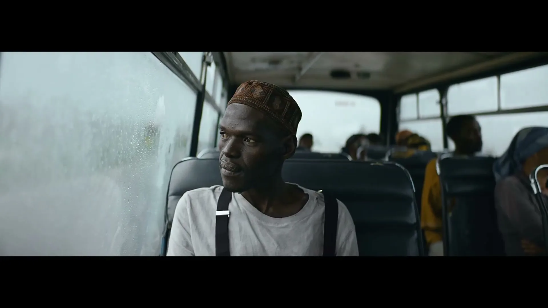 A man in a patterned cap and suspenders sits on a bus, looking out the window with a thoughtful expression.