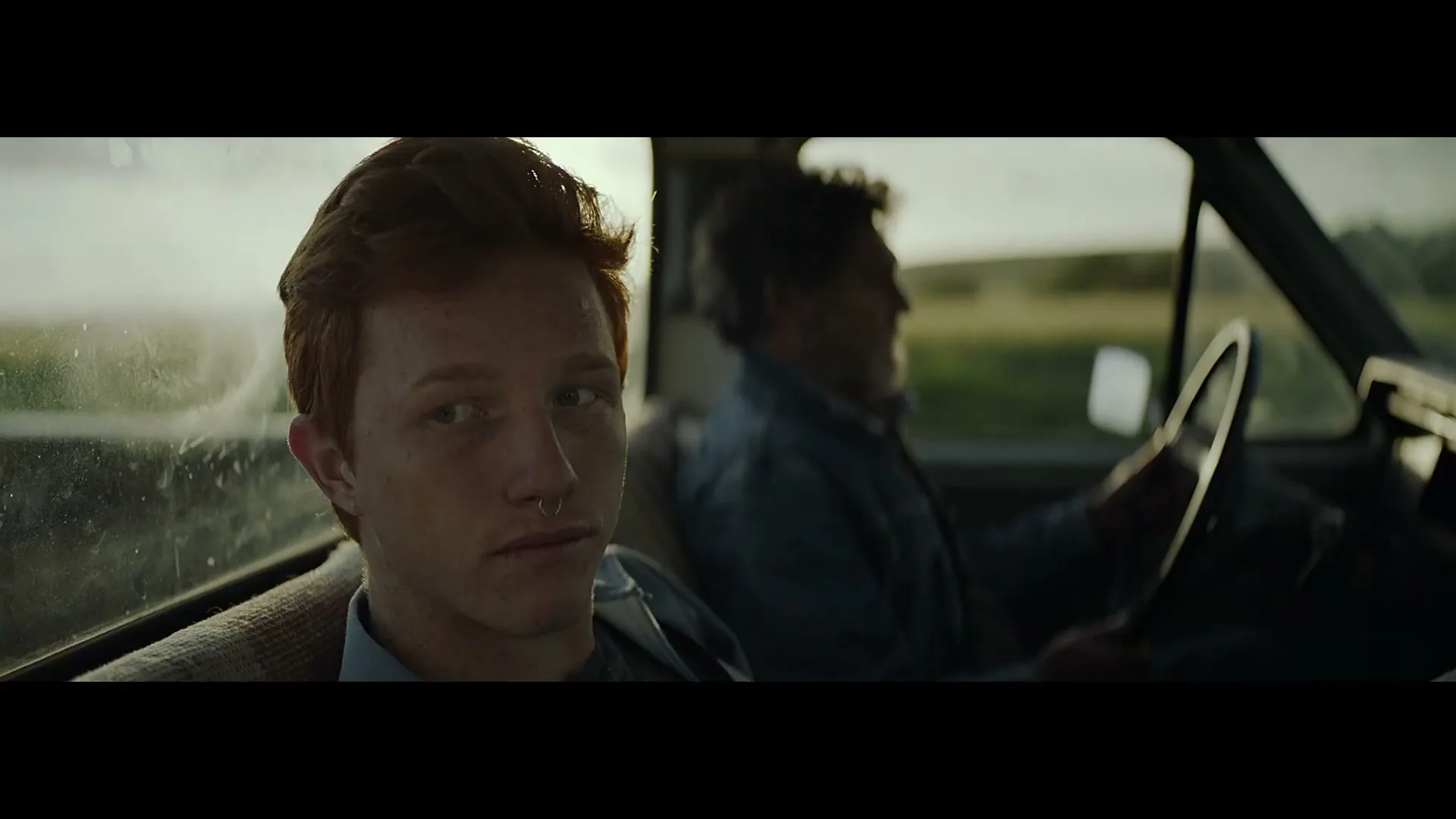 A young man with red hair and a nose ring sits in the passenger seat of a moving truck, looking forward, while an older man drives.