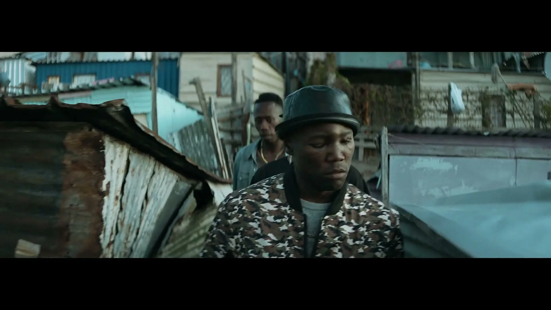 A young Black man in a camouflage jacket and black hat walks through a shantytown, looking around with a serious expression.