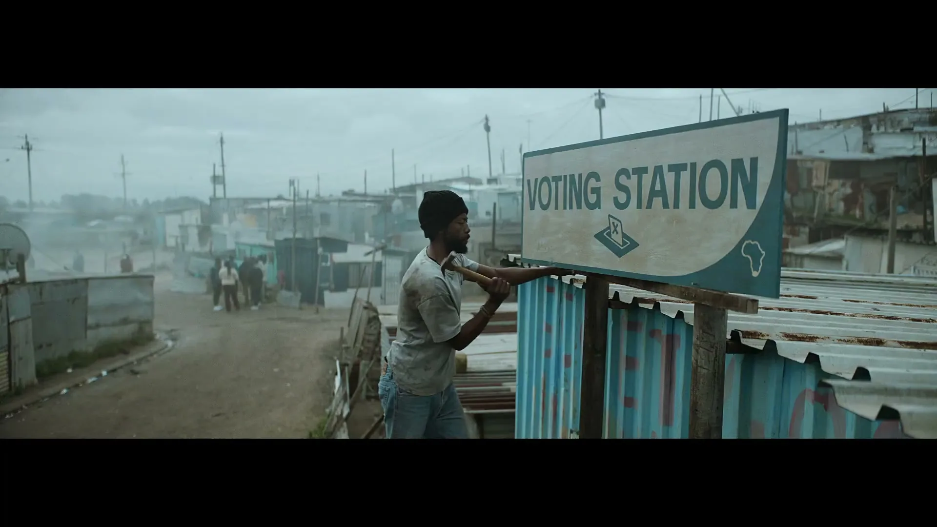 A man in a t-shirt and jeans hammers a "Voting Station" sign onto a corrugated metal fence in a dusty, crowded shantytown.