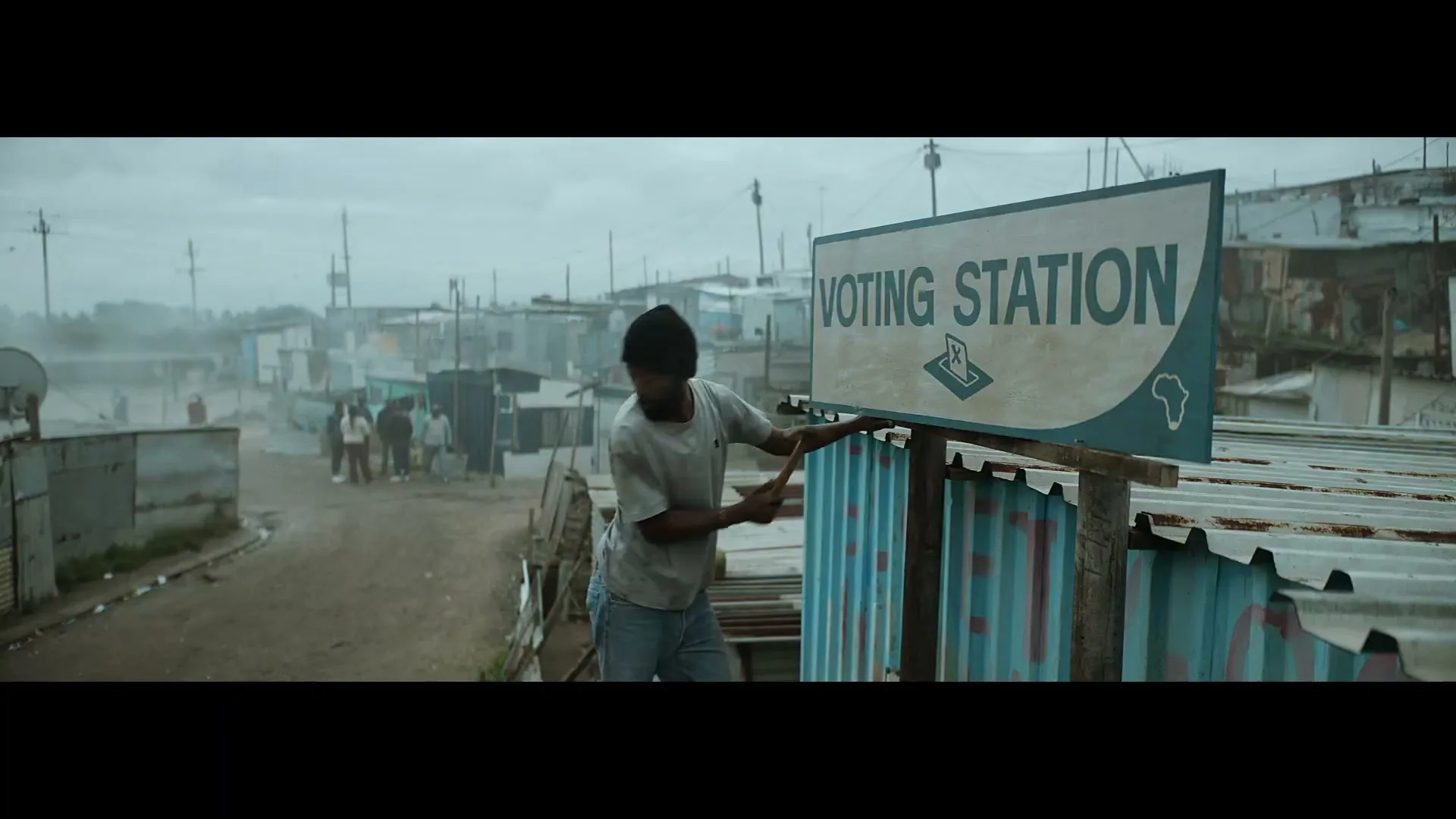 A man in a beanie and t-shirt hammers a "VOTING STATION" sign onto a corrugated metal fence in a dusty, crowded shantytown.