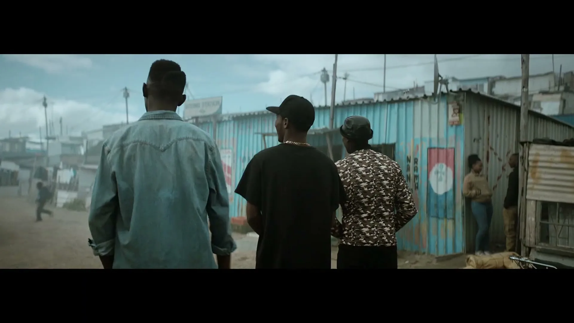 Three young men walk away from the camera down a dirt road in a crowded, impoverished neighborhood with corrugated metal shacks.