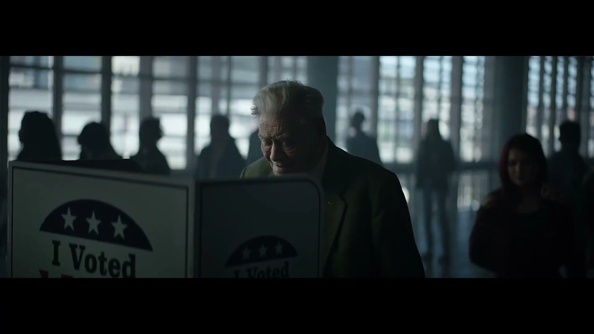 An elderly man with white hair and a green jacket stands at a voting booth, looking down intently, surrounded by blurred figures in a large, bright hall.