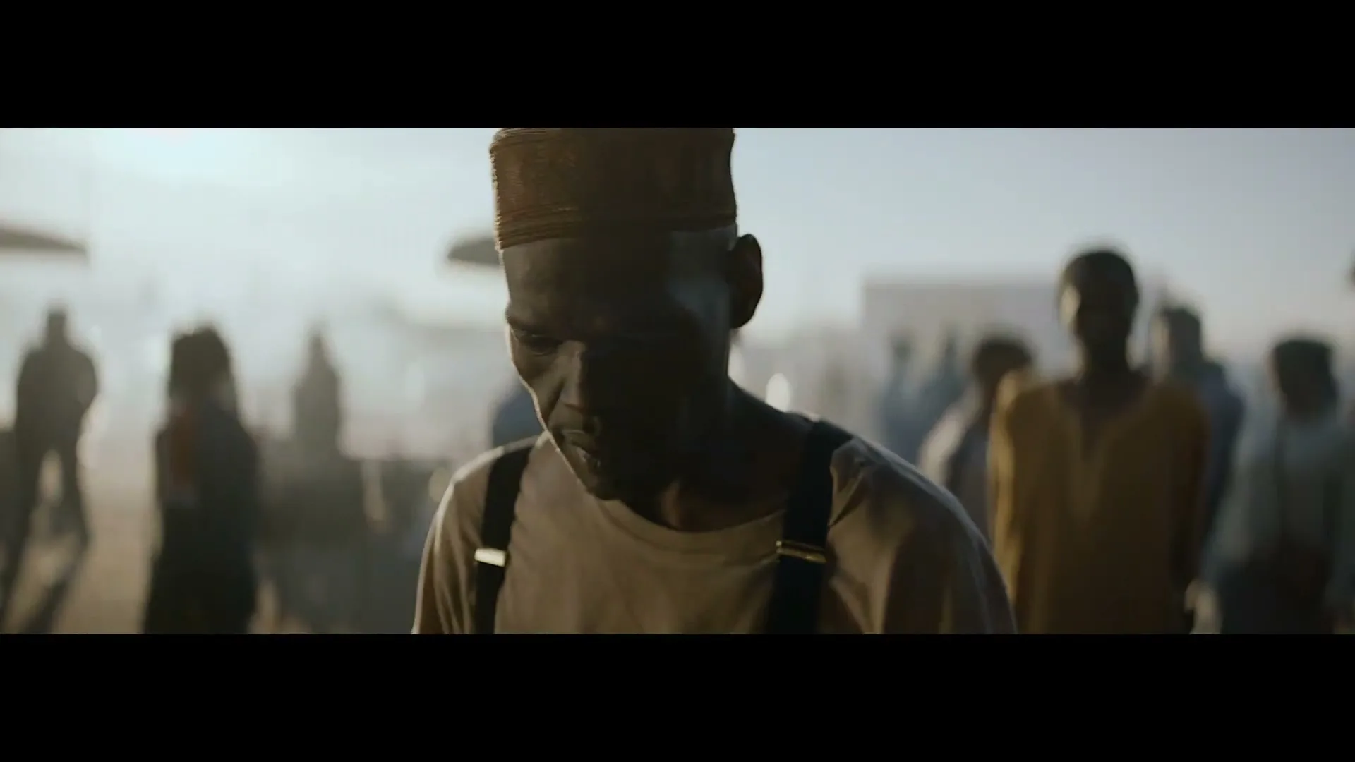 A man with dark skin and a cap looks down, surrounded by blurred people in a dusty, sunlit outdoor setting.
