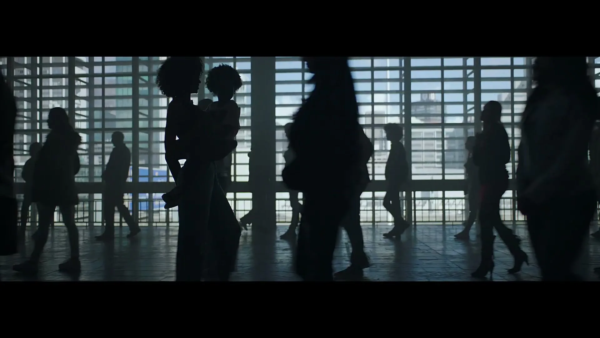 Silhouetted people, including a woman holding a child, walk past large windows in a modern building, with a city visible outside.