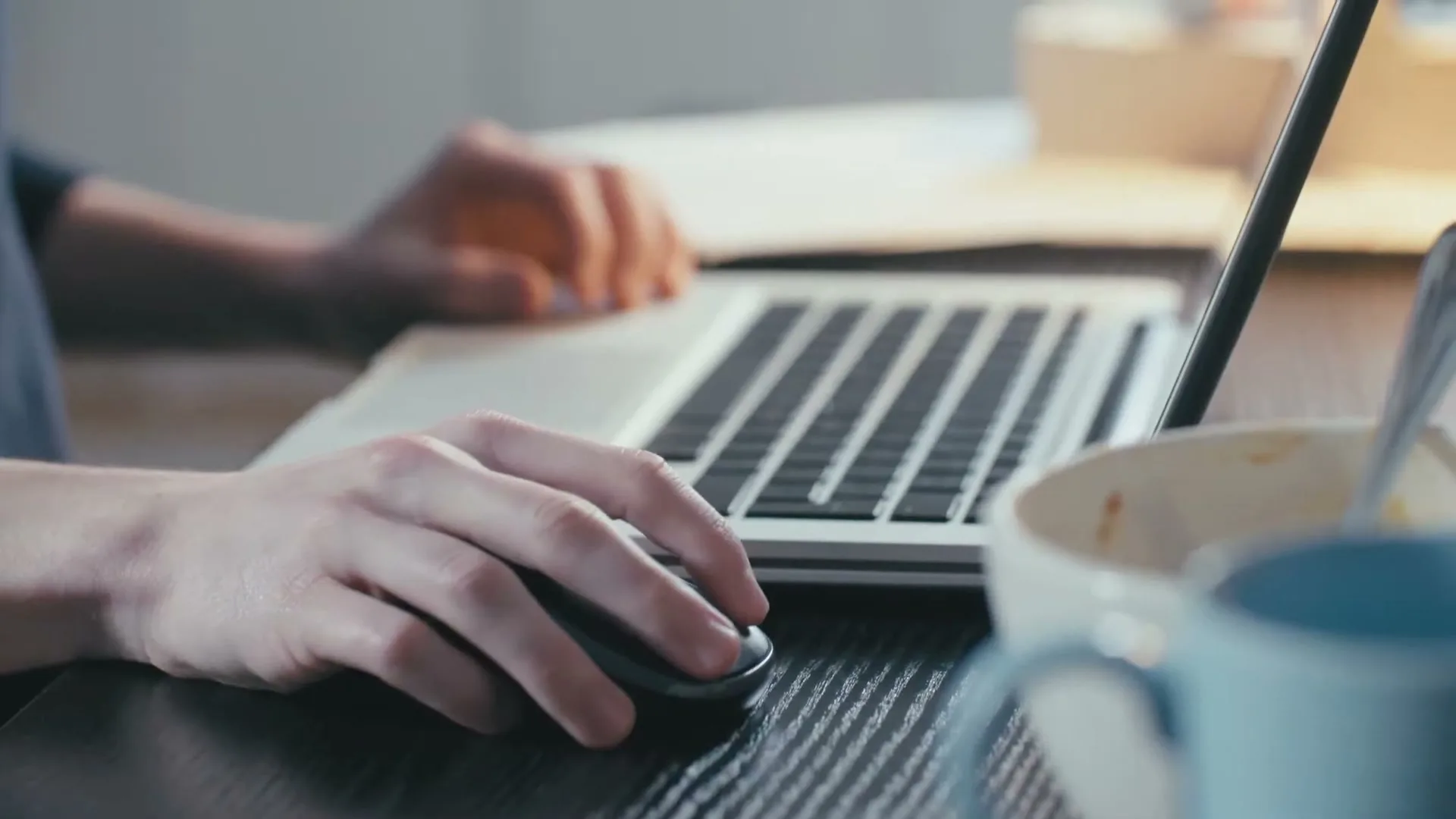 A person's hands are seen using a laptop and mouse on a wooden desk, with a bowl and mug nearby.