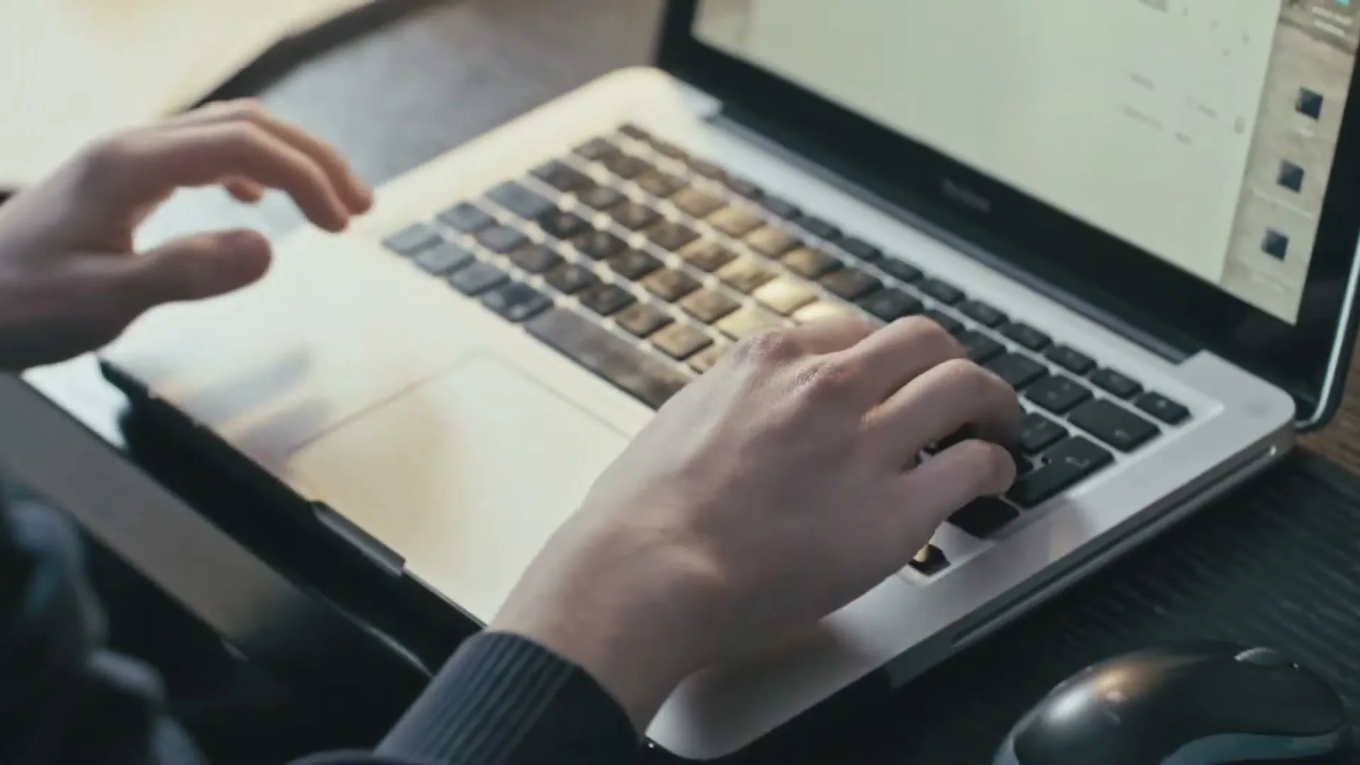 A person's hands are seen typing on a silver laptop keyboard, with a computer mouse visible on the right side of the frame, on a dark wooden desk.