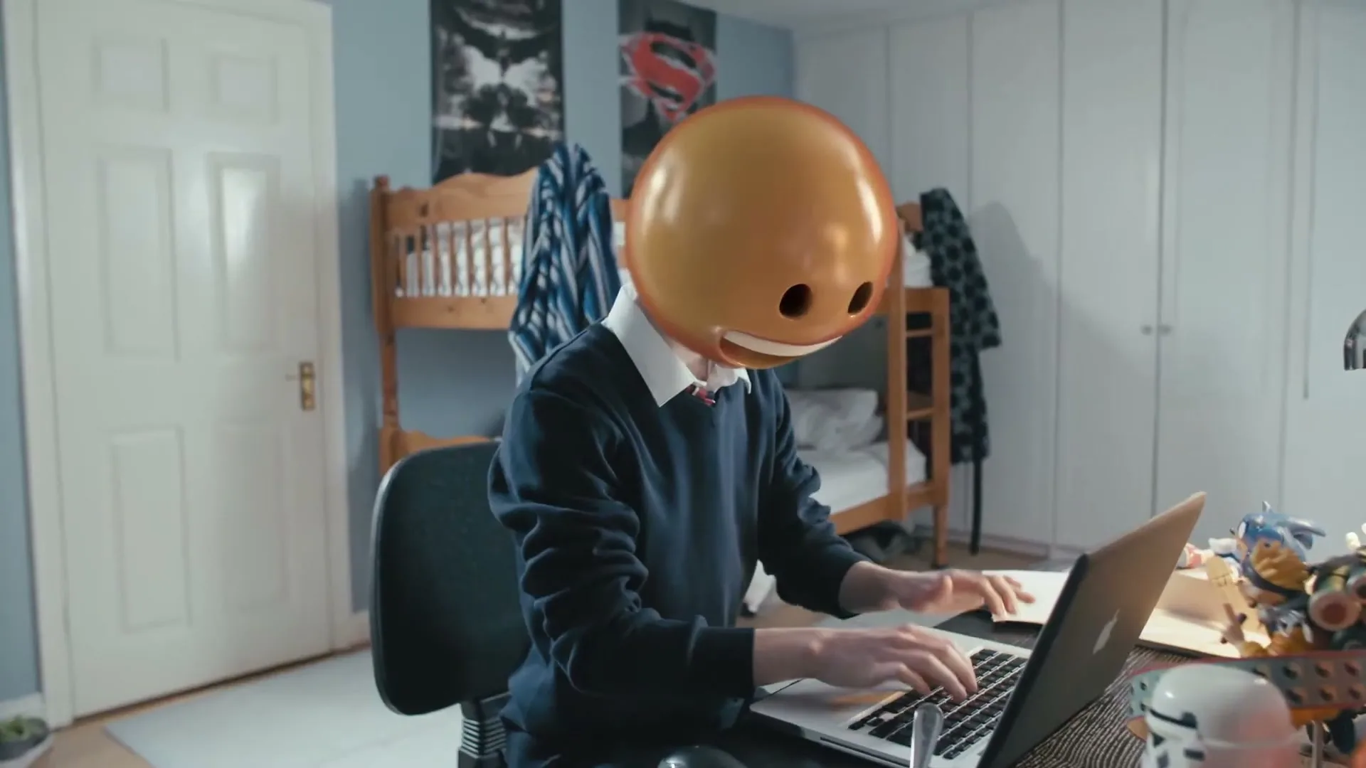 A teen with an emoji mask sits at a desk, typing on a laptop in a bedroom.
