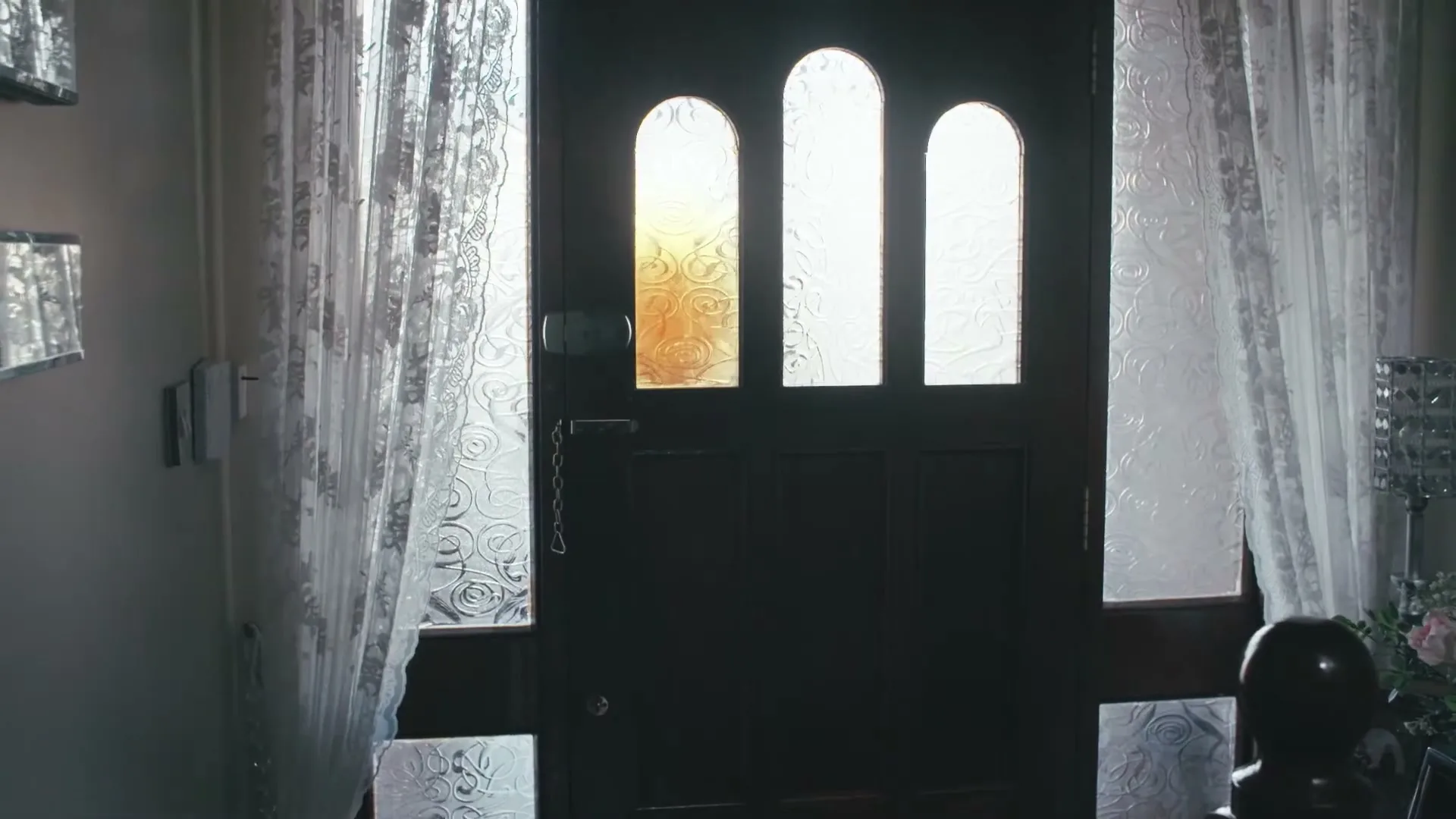 A dark wooden door with three arched glass panels, flanked by sheer white curtains, is seen from inside a house.