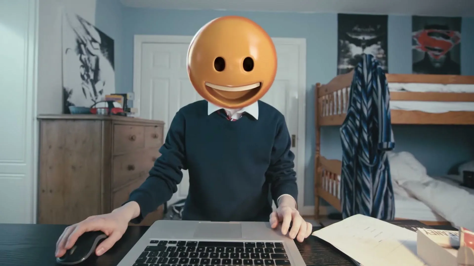 A teen with an emoji head sits at a desk, using a laptop and mouse in a bedroom.