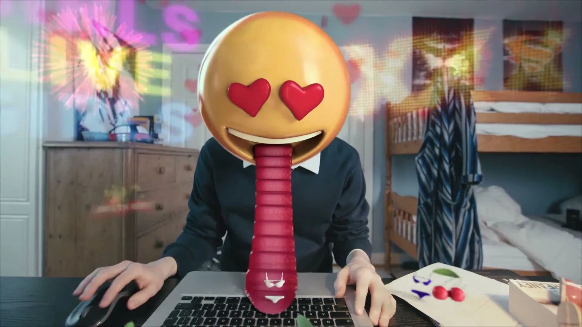 A person with an emoji head sits at a desk with a laptop, as animated hearts and suggestive imagery appear around them in a bedroom.