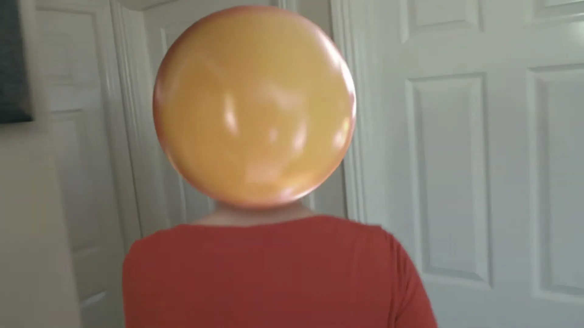 A person with a large, round, orange emoji-like head walks away from the camera in a house.