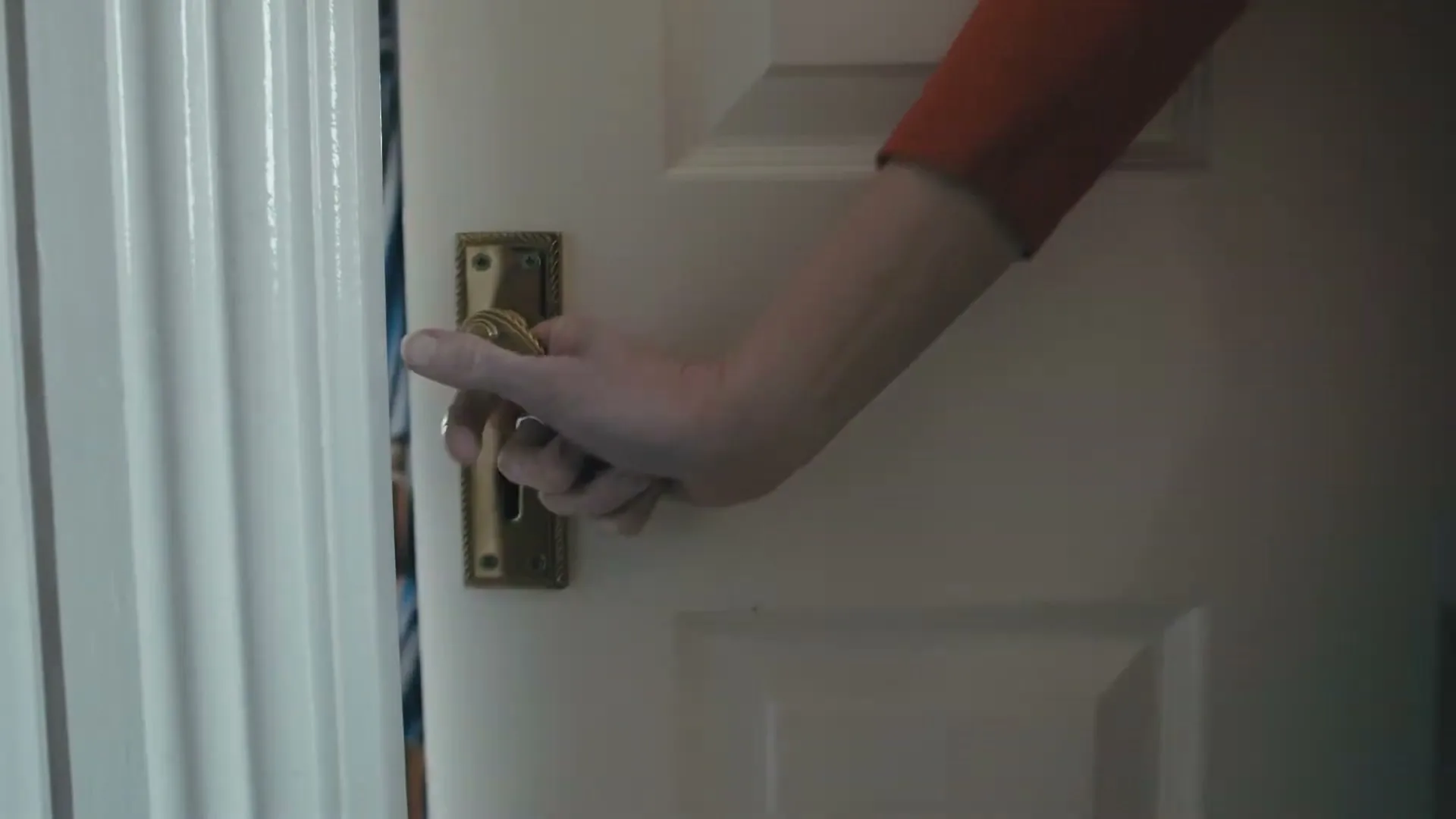 A person's hand reaches for a brass doorknob on a white door, turning it and opening the door to reveal a room beyond.
