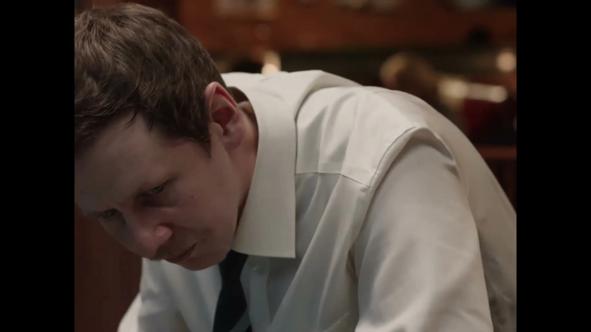 A man in his 30s, wearing a white shirt and tie, looks down with a serious expression in an interior setting.