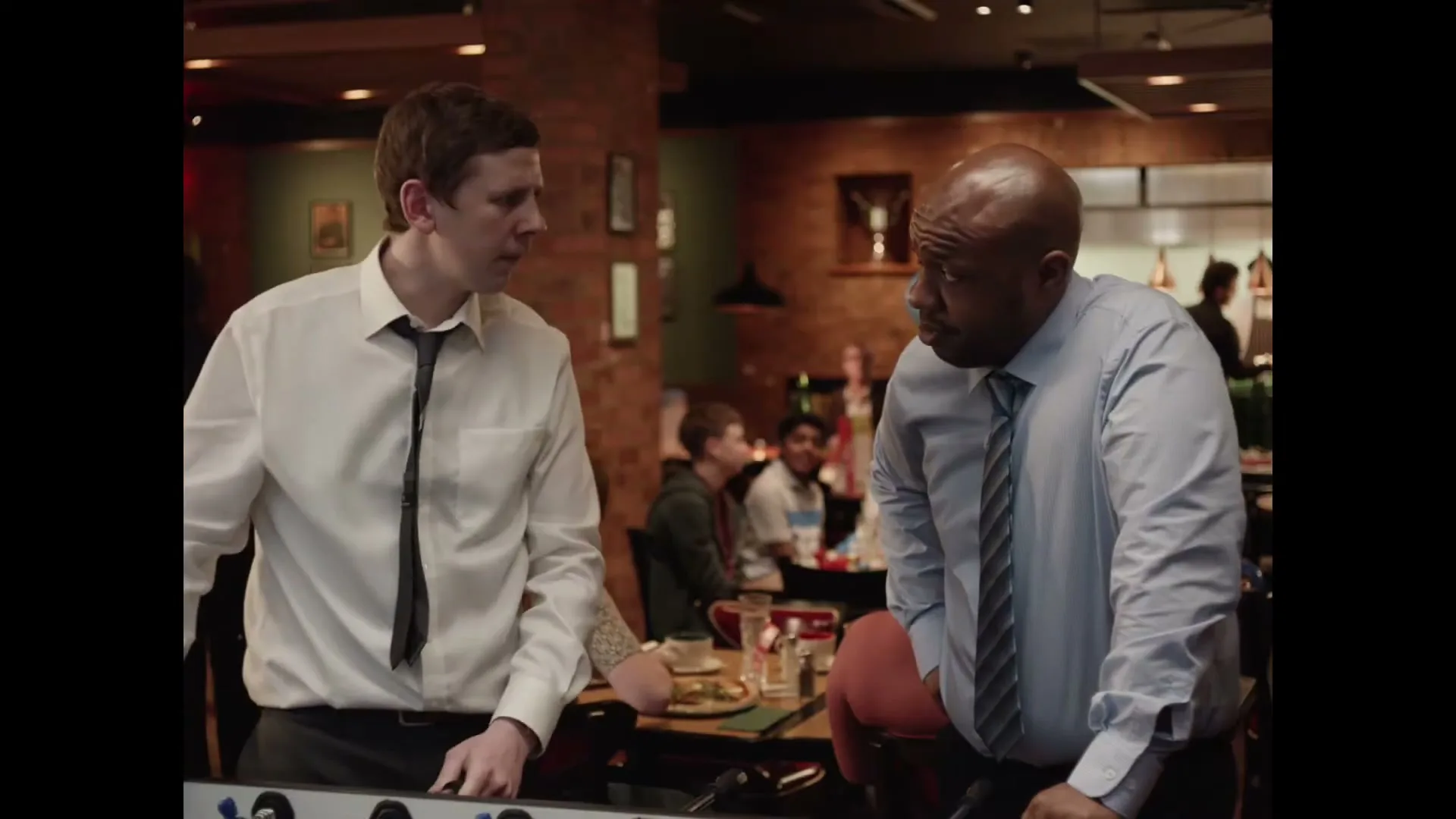 Two men in business casual attire play foosball in a restaurant, with one man looking intently at the other.