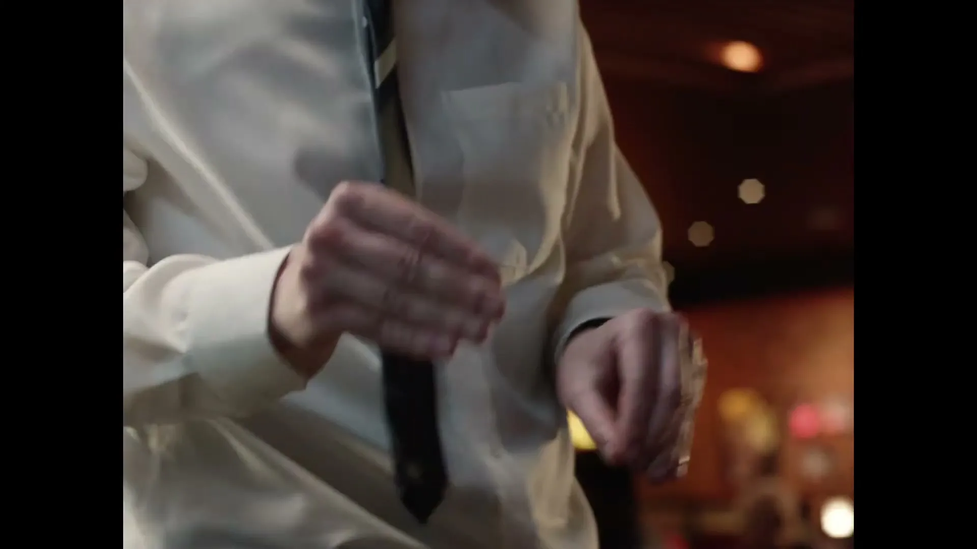 A man in a white shirt and tie gestures with his hands in a dimly lit interior setting.