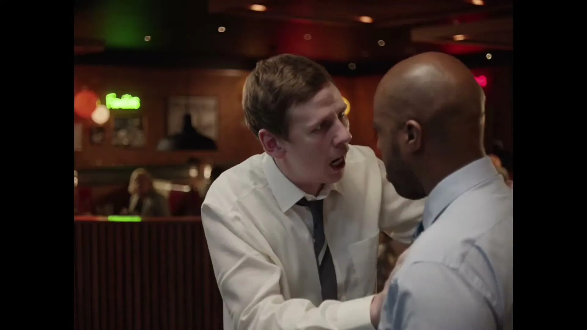 A white man in a white shirt and tie aggressively talks to a Black man in a blue shirt in a dimly lit restaurant.