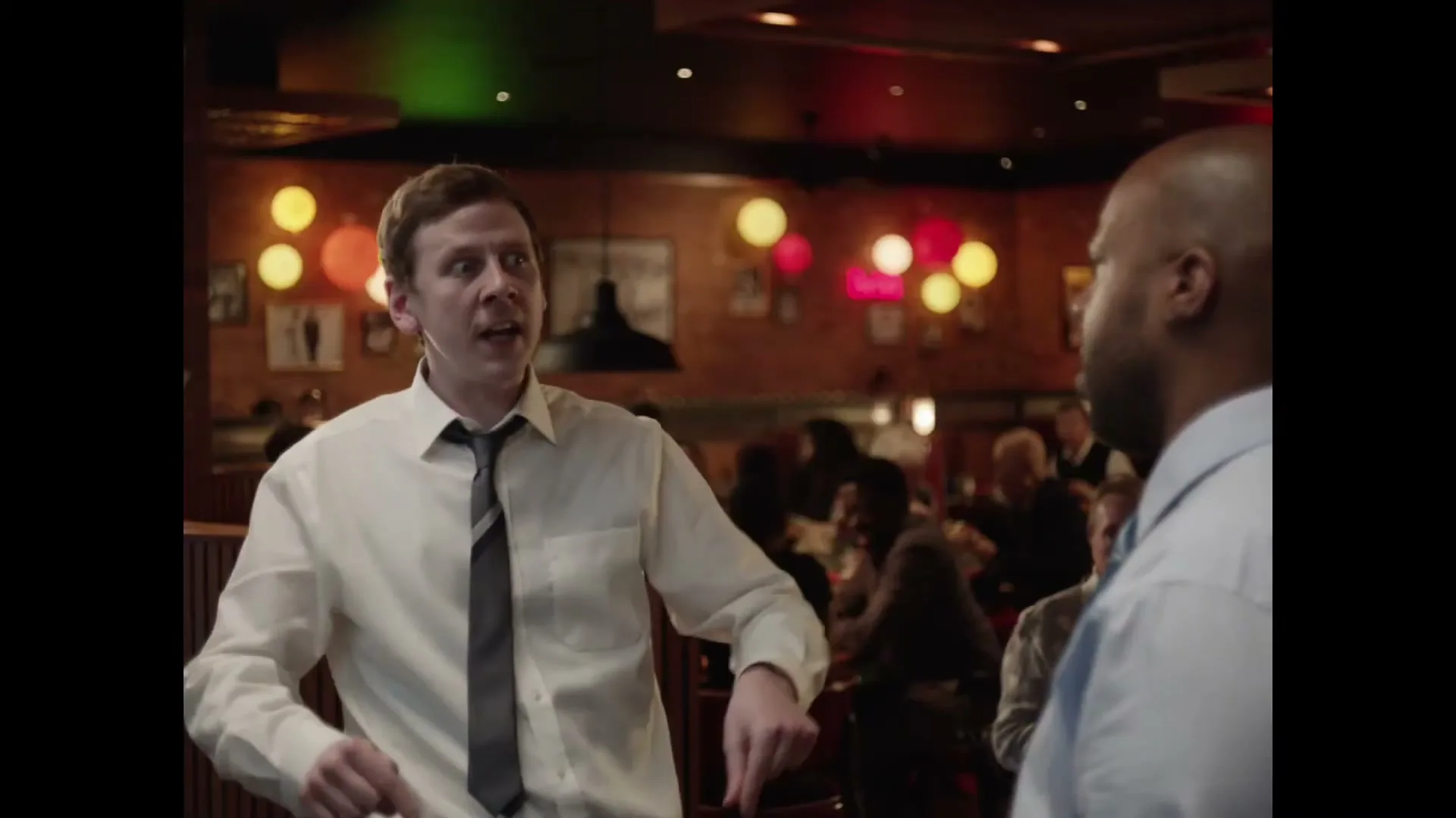 Two men in business casual attire talk in a busy restaurant or bar with colorful lights in the background.