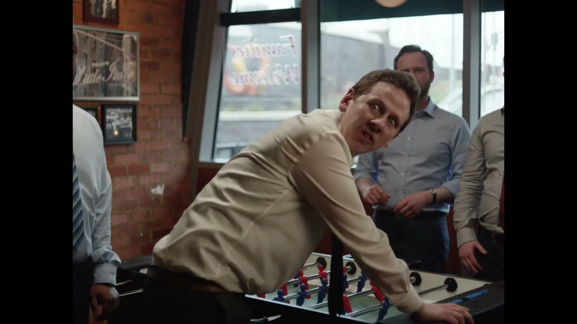 A man in a beige shirt leans over a foosball table, looking surprised, while other men in business casual attire stand around him in an office breakroom.