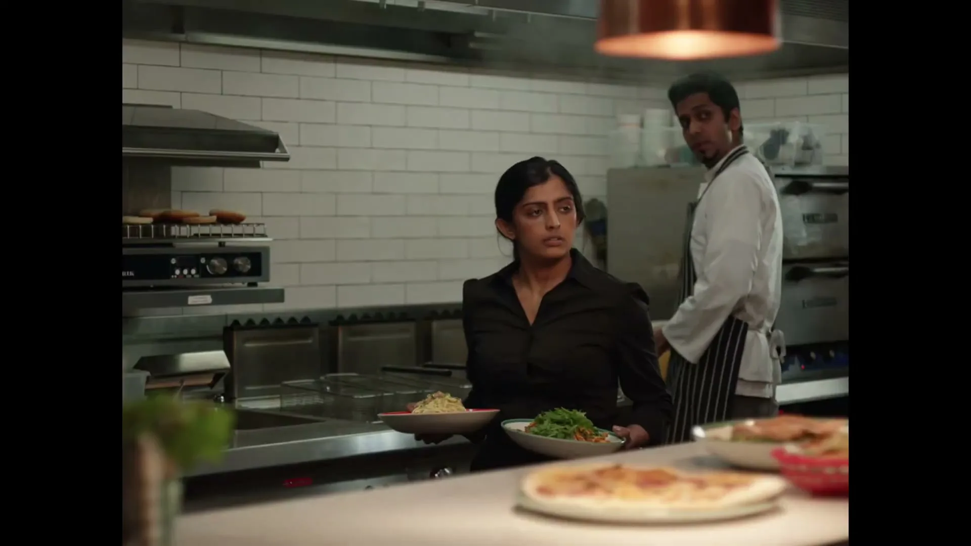 A female server in a black shirt holds two plates of food, looking off-camera in a busy restaurant kitchen, with a male chef in the background.