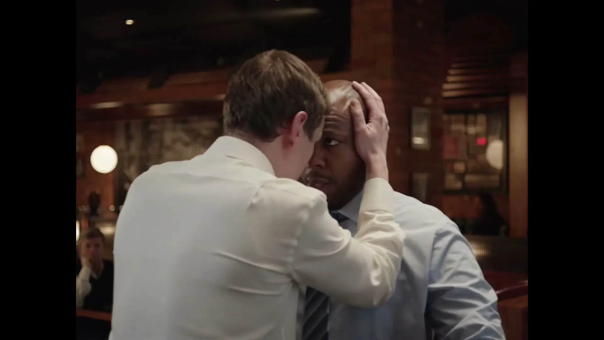 A white man in a white shirt leans in close to a Black man in a blue shirt and tie, holding his head in a dimly lit bar.