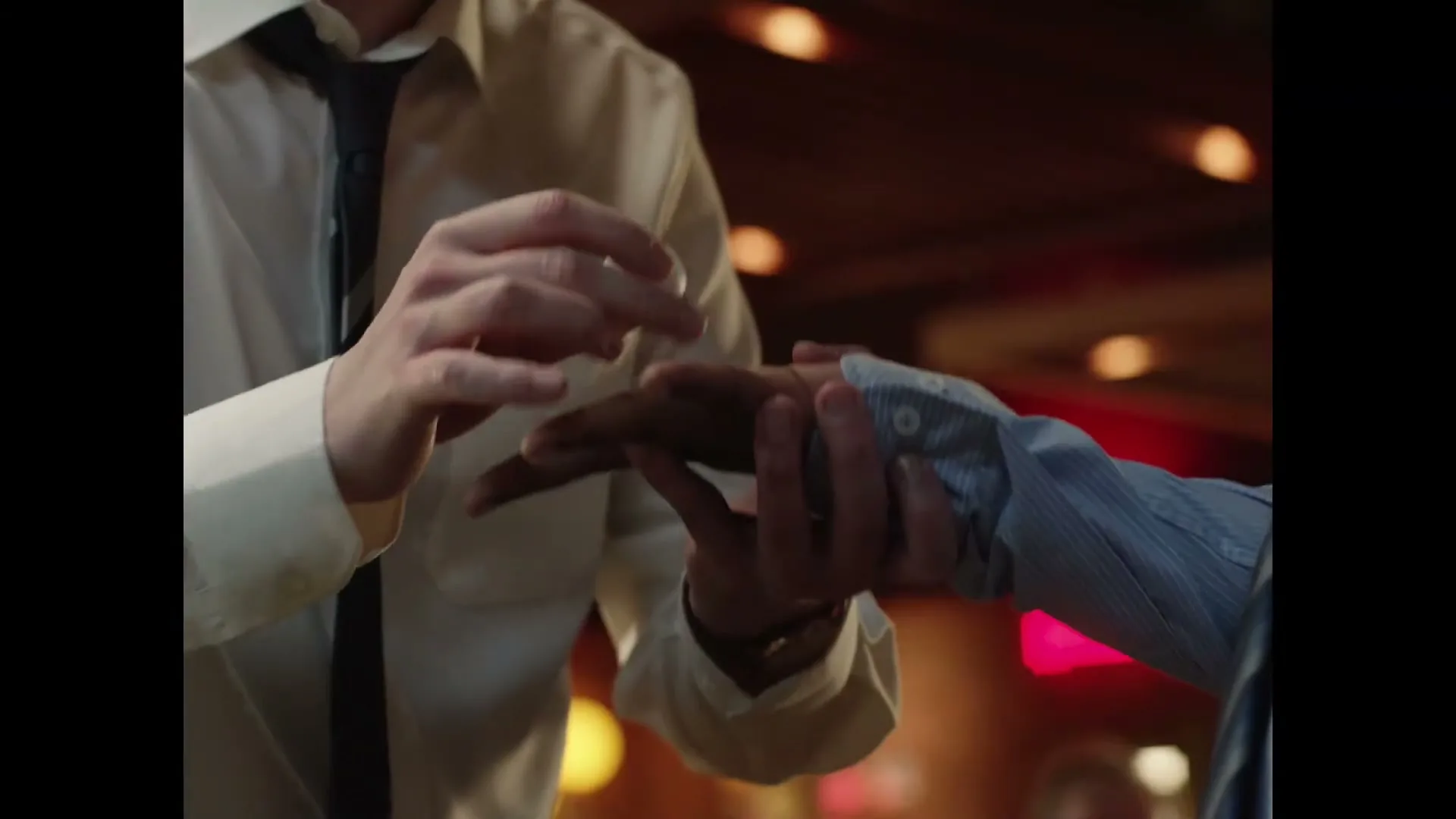A man in a white shirt and tie performs a magic trick, making a coin appear in another person's hand in a dimly lit interior.