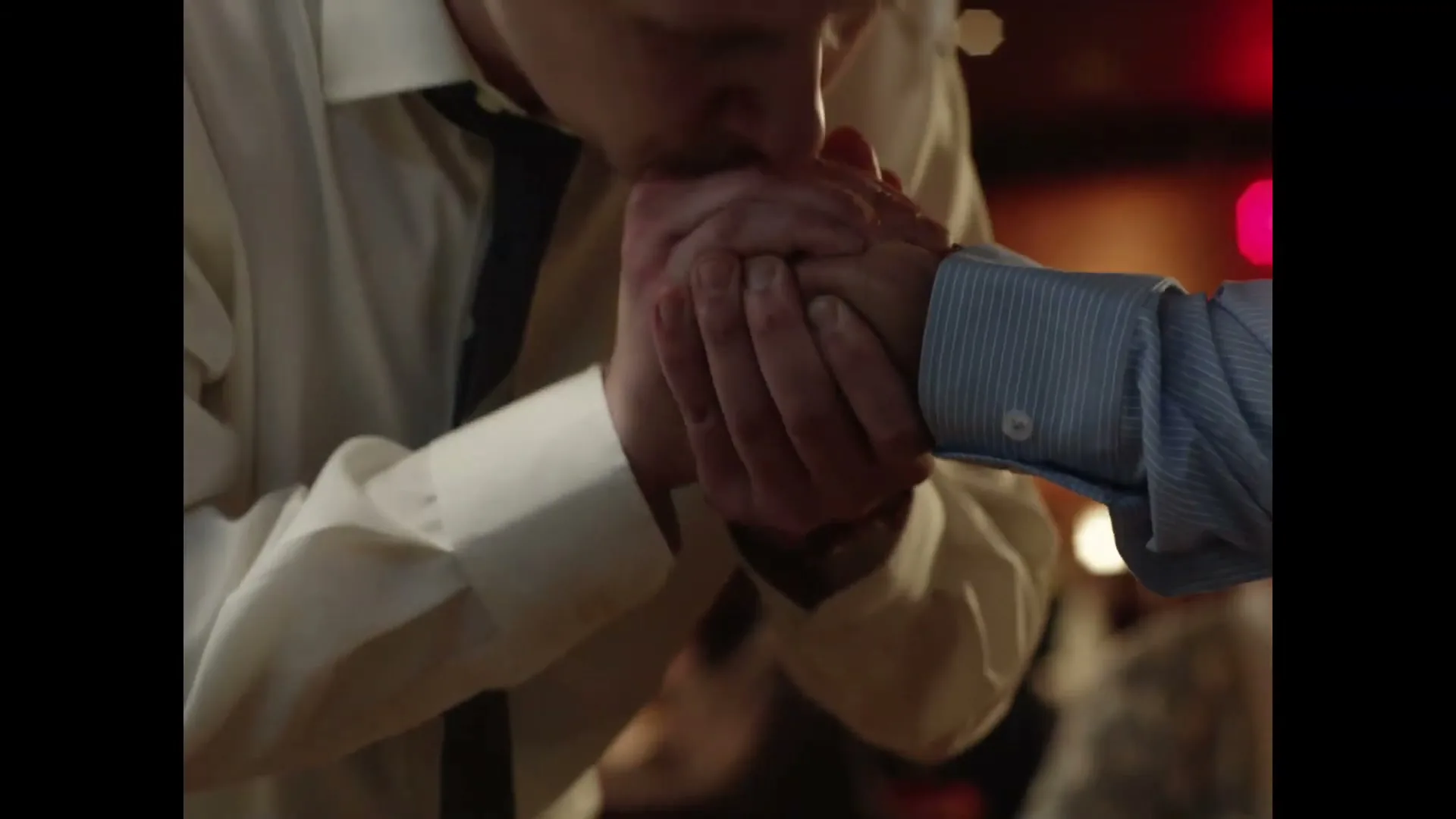 A man in a white shirt and tie kisses the hand of another person in a blue striped shirt, showing affection in a dimly lit interior.