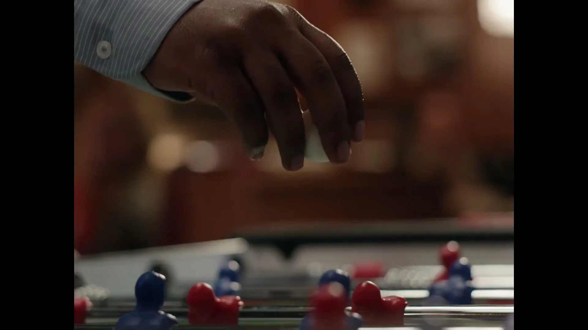 A person's hand drops a white foosball into the game, with red and blue players visible on the table.