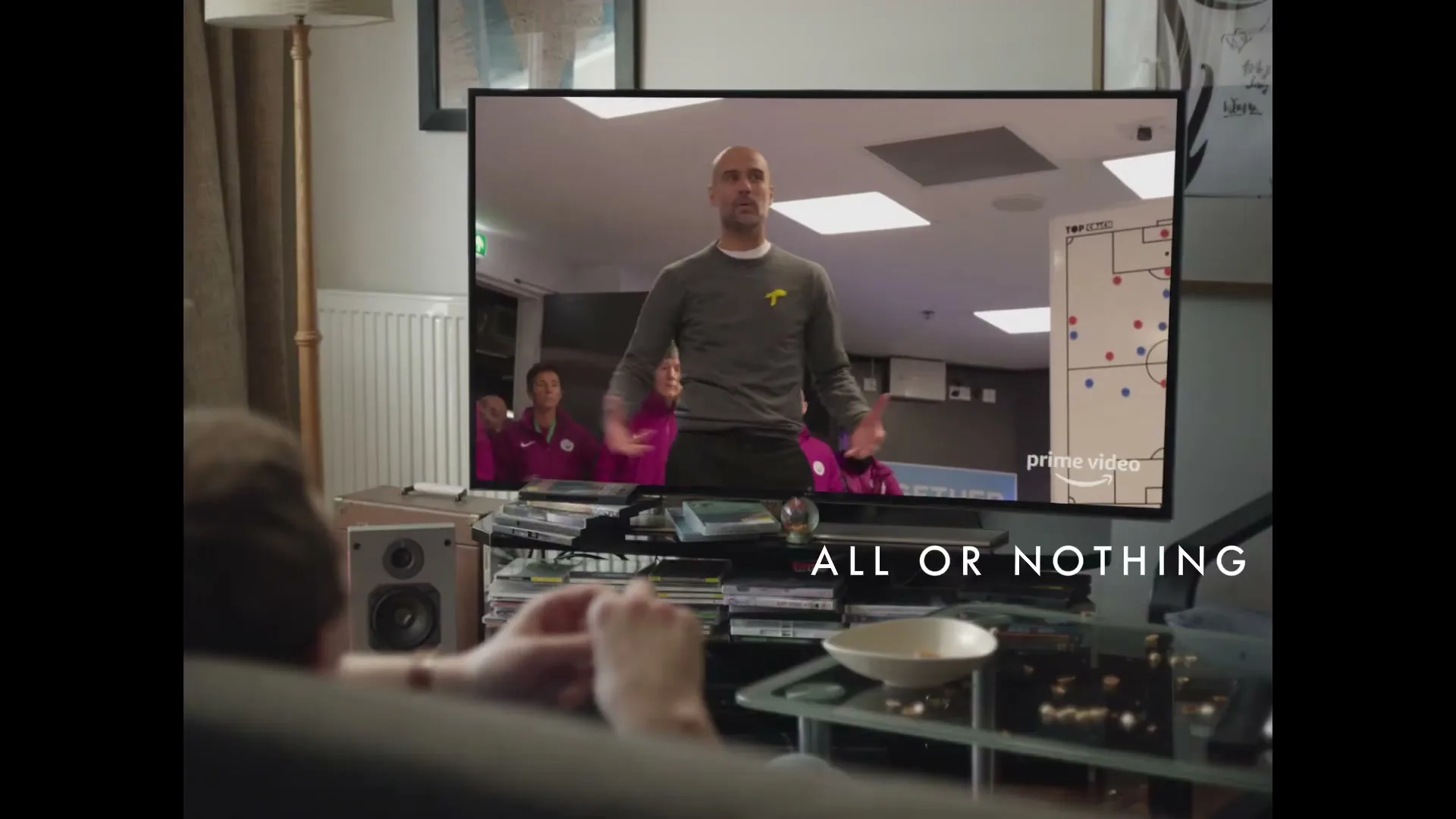 A person watches a football coach on a TV screen in a living room, with "ALL OR NOTHING" text on screen.