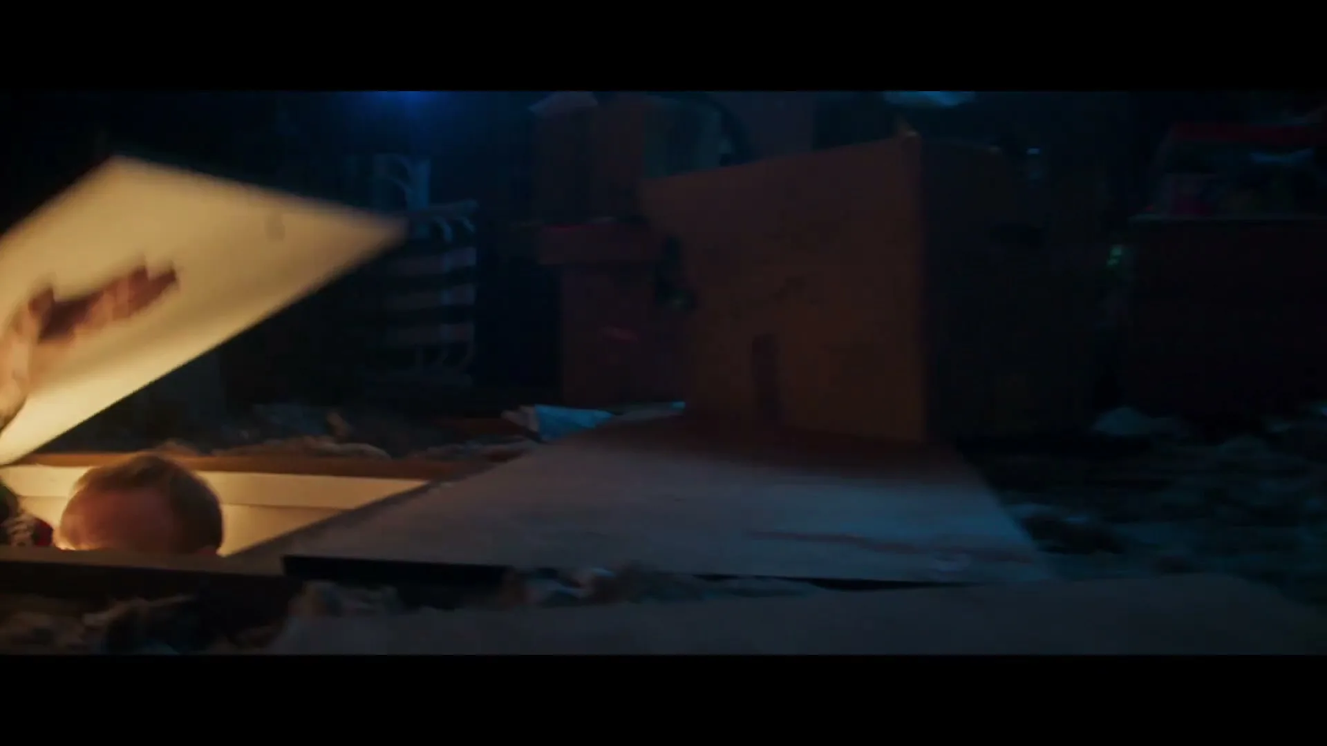 A man in a festive sweater emerges from a trapdoor in a dark, dusty attic, smiling as he pulls out a box labeled "Christmas Lights."