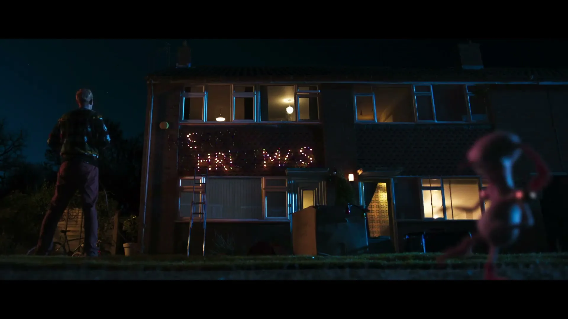 A man in a festive sweater watches as a small, alien-like creature removes letters from a "Merry Christmas" light display on a house at night.