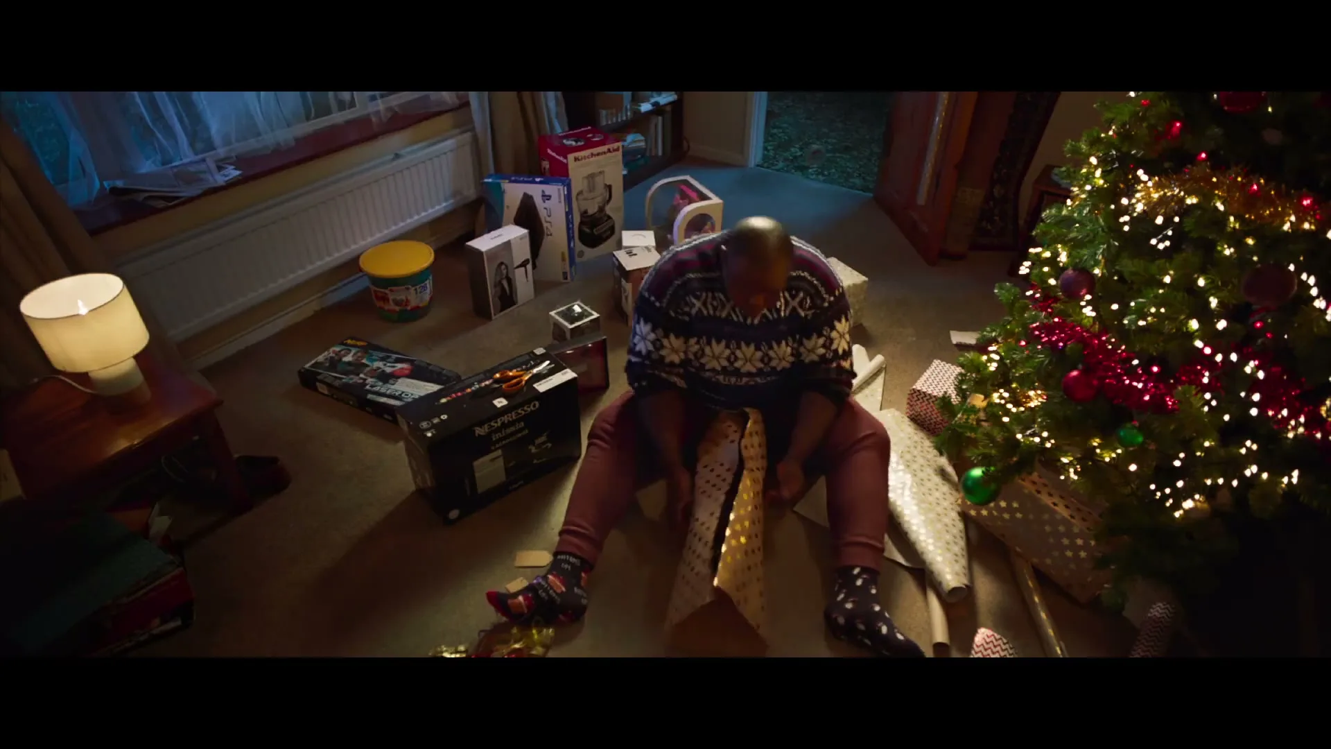 A man in a Christmas sweater sits on the floor, struggling to wrap a gift next to a decorated Christmas tree in a living room.