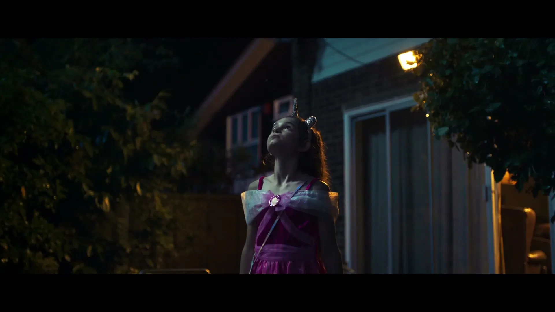 A young girl in a unicorn costume looks up at the sky in a suburban backyard as the lighting shifts from day to night.