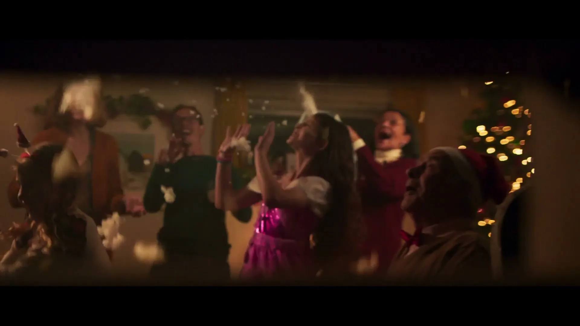 A group of diverse people of various ages are seen through a mail slot, laughing and throwing fake snow at a Christmas party.