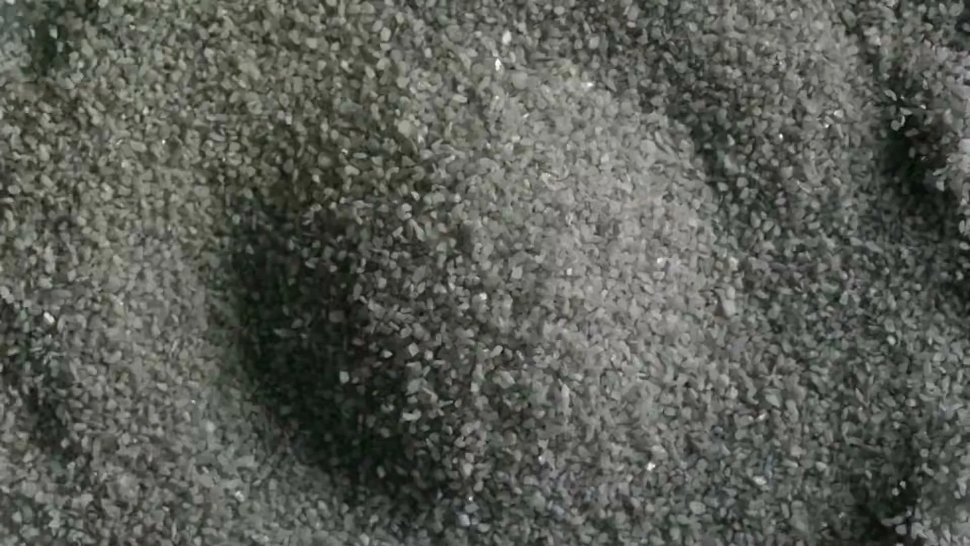 A top-down, extreme close-up shot of small, dark grey gravel or sand, with subtle light reflections, creating a textured, abstract visual.