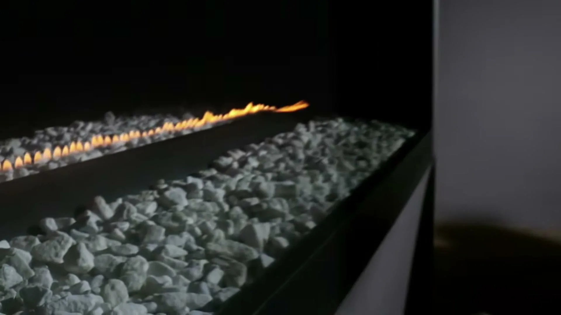 A modern fireplace with white stones and a line of flames igniting, in a dimly lit room.