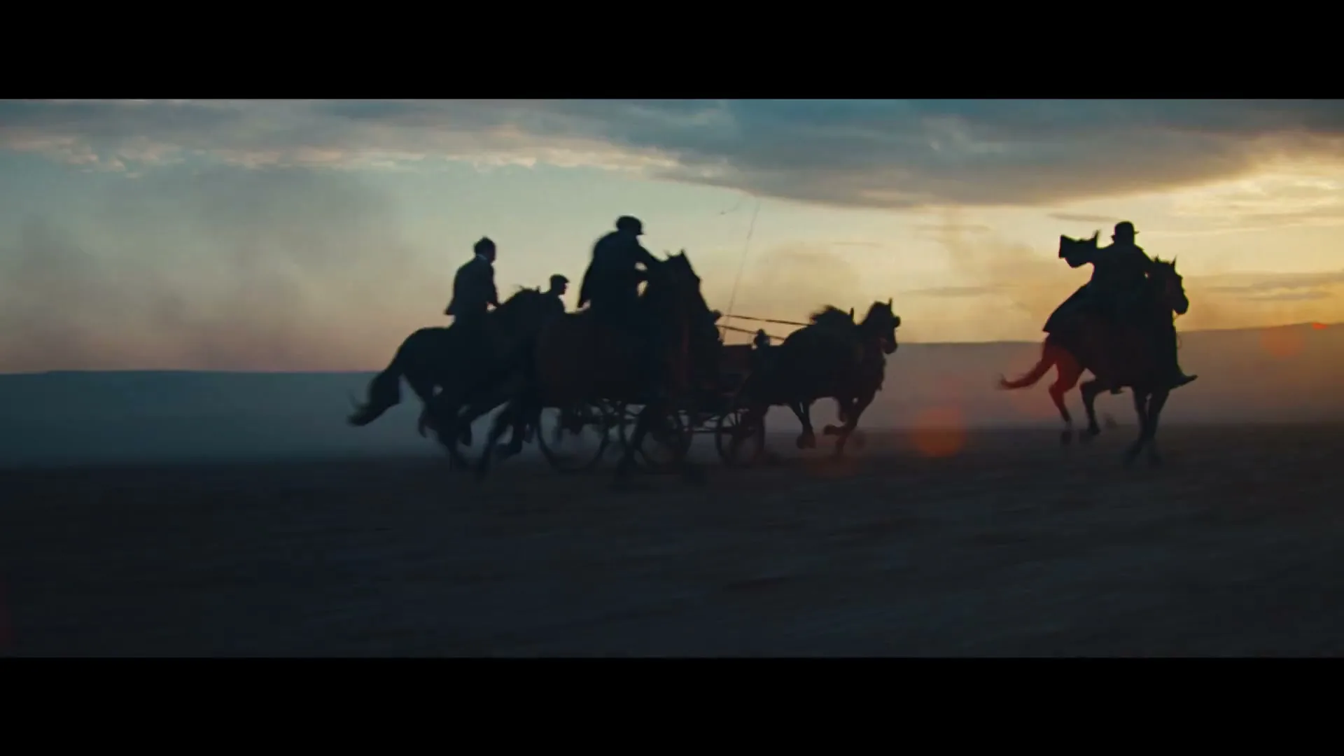 Silhouetted figures on horseback and a horse-drawn carriage gallop across a vast, dusty plain at sunset.
