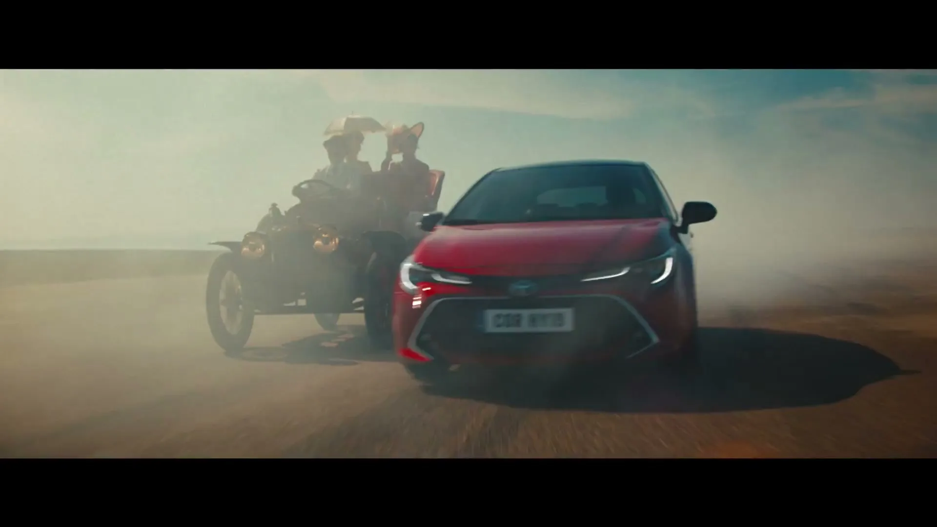 A red Toyota car drives on a dusty road, passing an old-fashioned car with two people wearing fox masks.