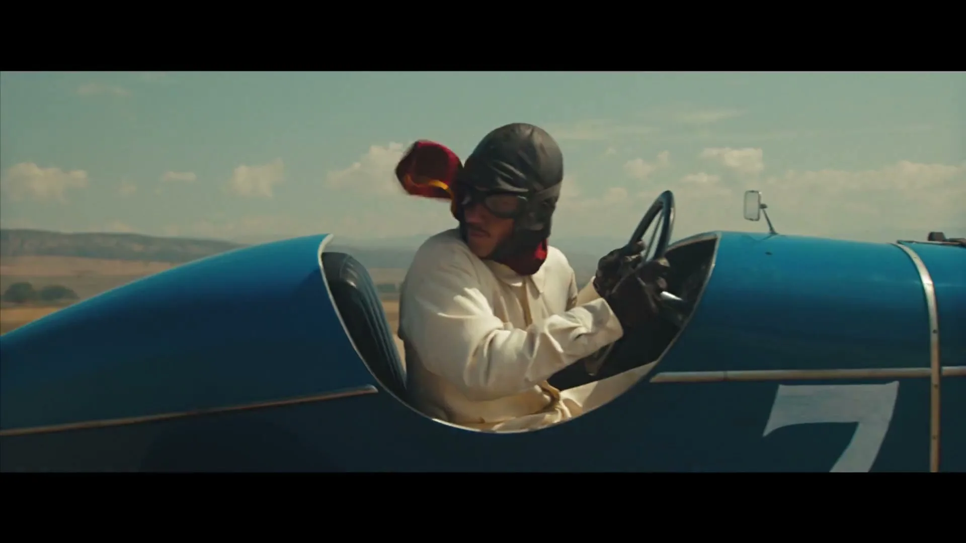 A male race car driver in a vintage blue car with a red scarf and helmet drives on a dirt track under a blue sky.