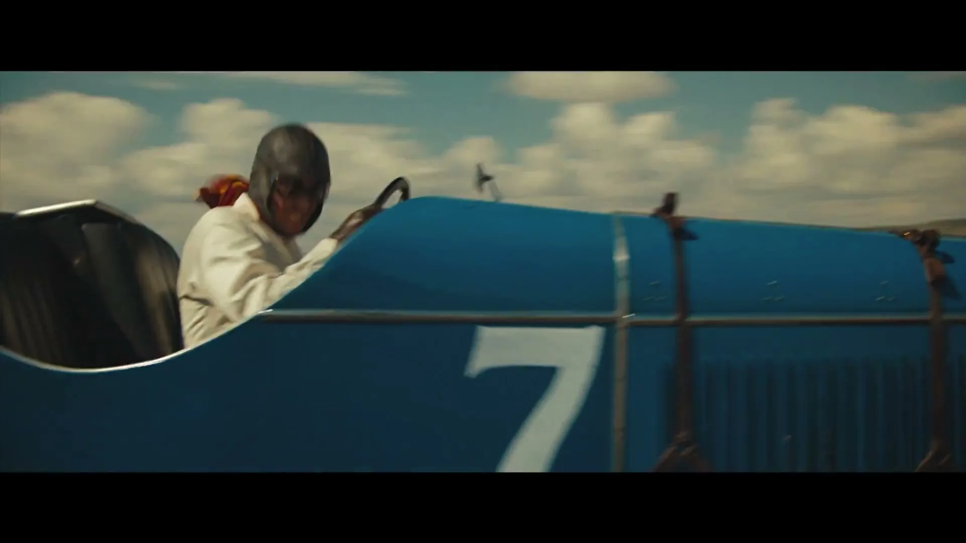 A male race car driver in a vintage blue car with the number 7 on its side, drives on a sunny day.