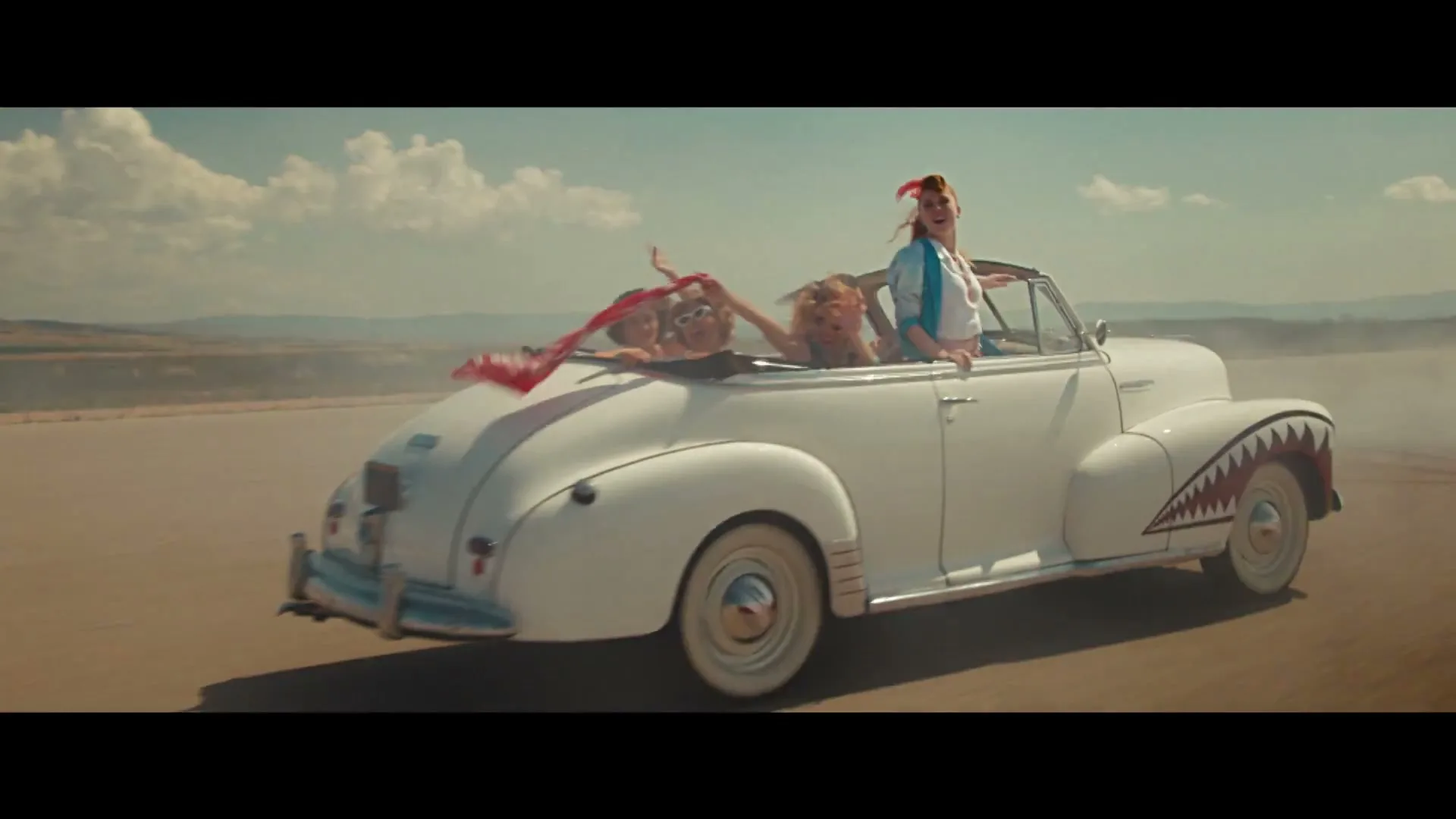 Four women in retro outfits ride in a white vintage convertible with shark teeth painted on the side, driving on a dusty road under a blue sky.