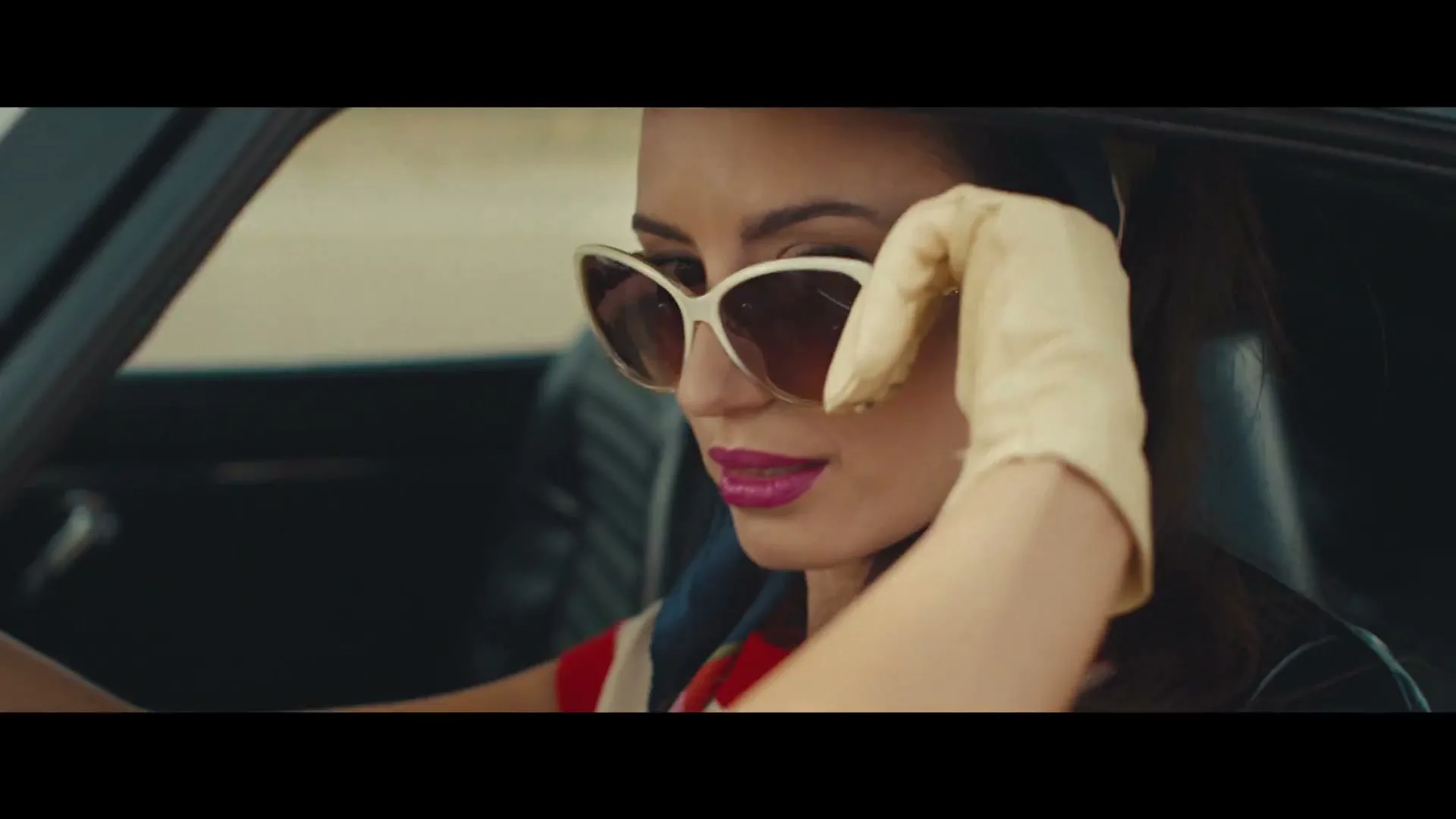 A woman in a vintage car, wearing sunglasses and gloves, adjusts her sunglasses while looking to the side.
