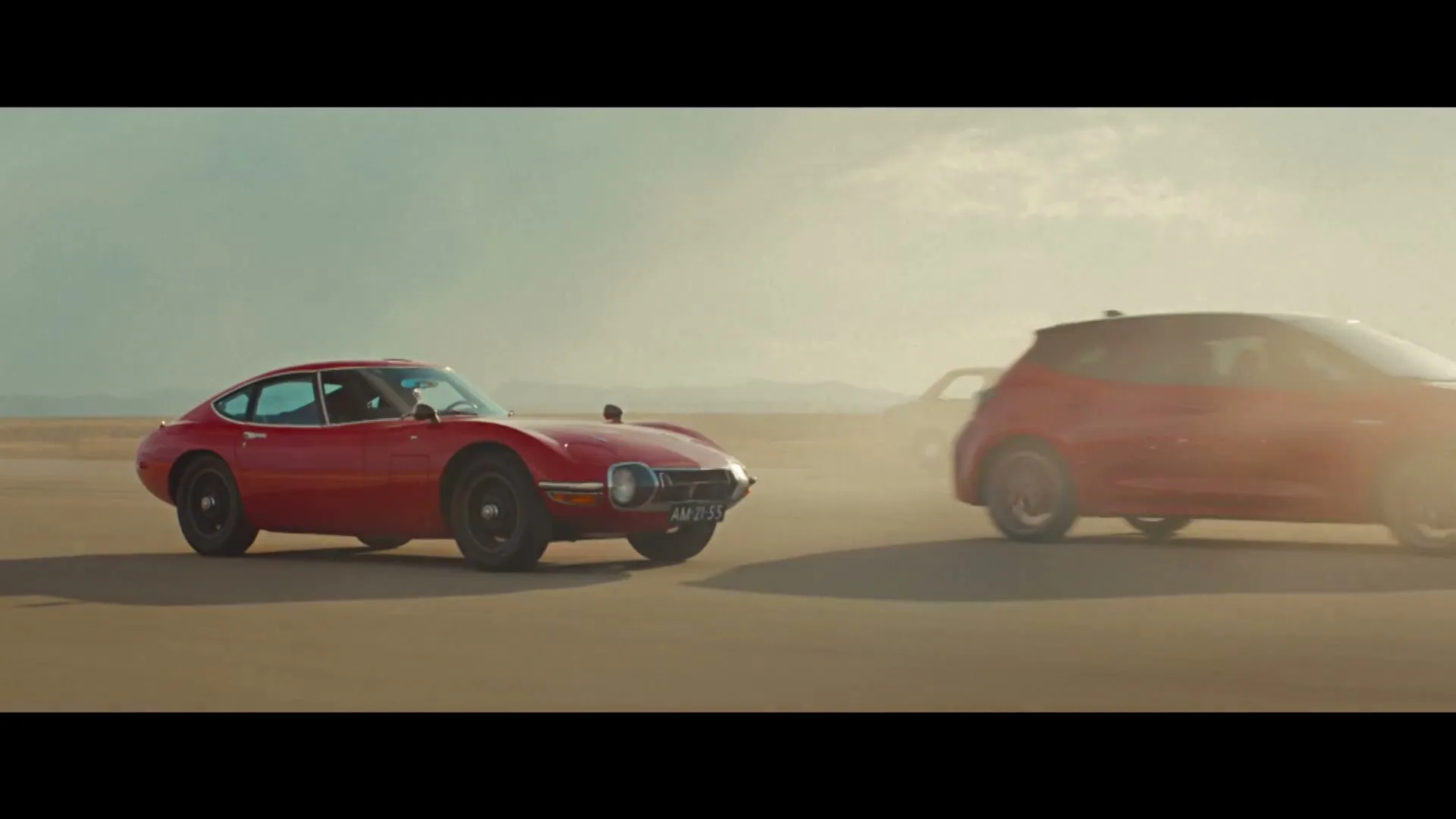A red vintage Toyota 2000GT and a red modern Toyota Corolla hatchback drive side-by-side on a dusty desert road under a cloudy sky.