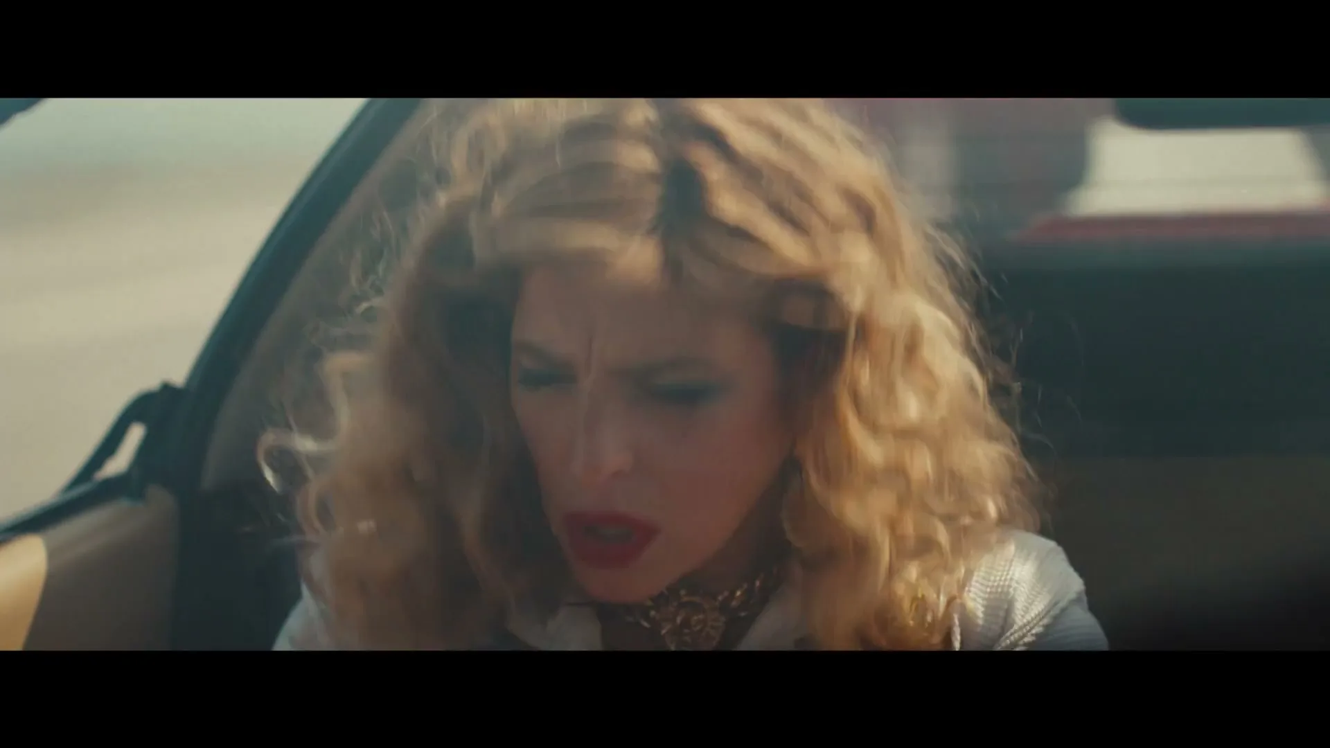 A woman with curly blonde hair and red lipstick, wearing a white jacket and gold jewelry, looks around anxiously in a car.