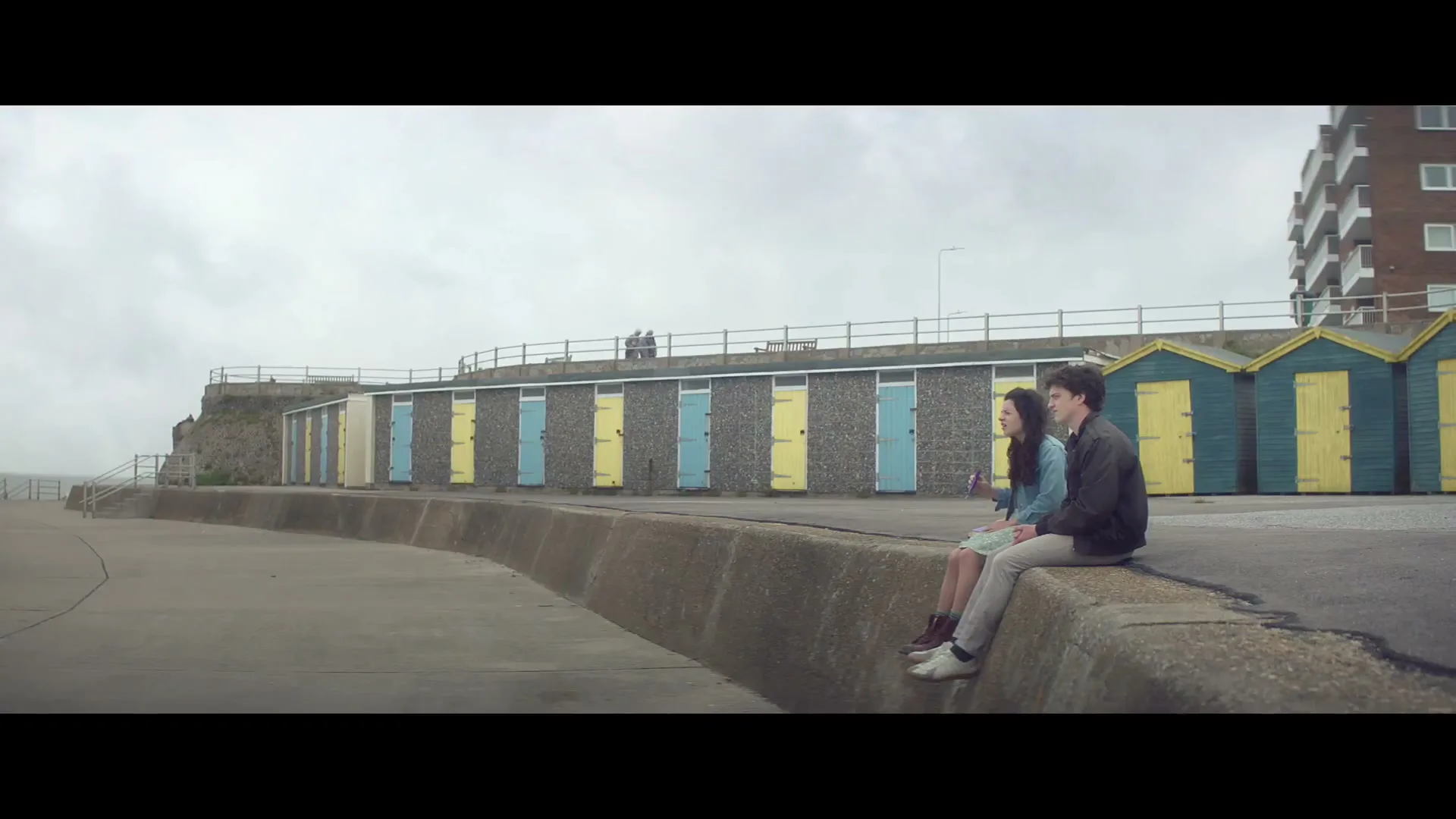 A young couple sits on a concrete wall, looking out at the sea on a cloudy day, with colorful beach huts in the background.