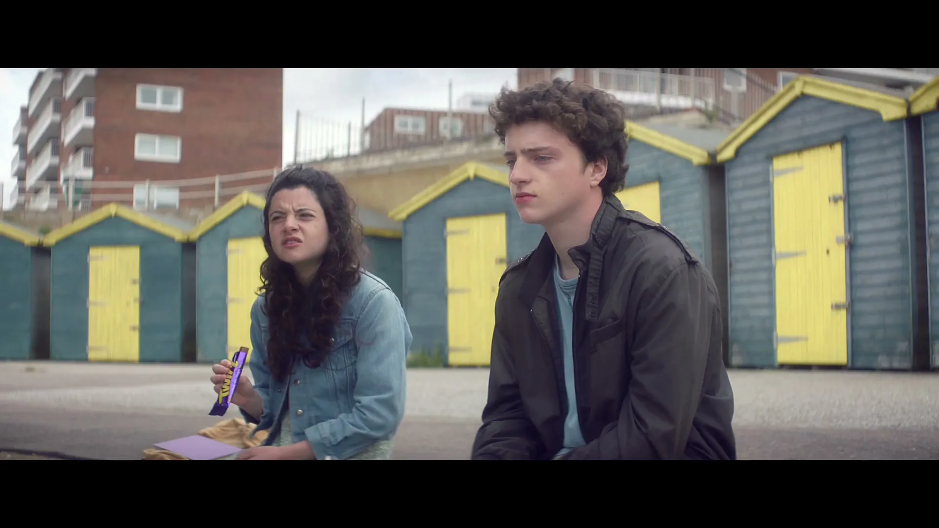 A young woman and man sit on a concrete wall, with yellow and blue beach huts behind them, as the woman eats a chocolate bar.