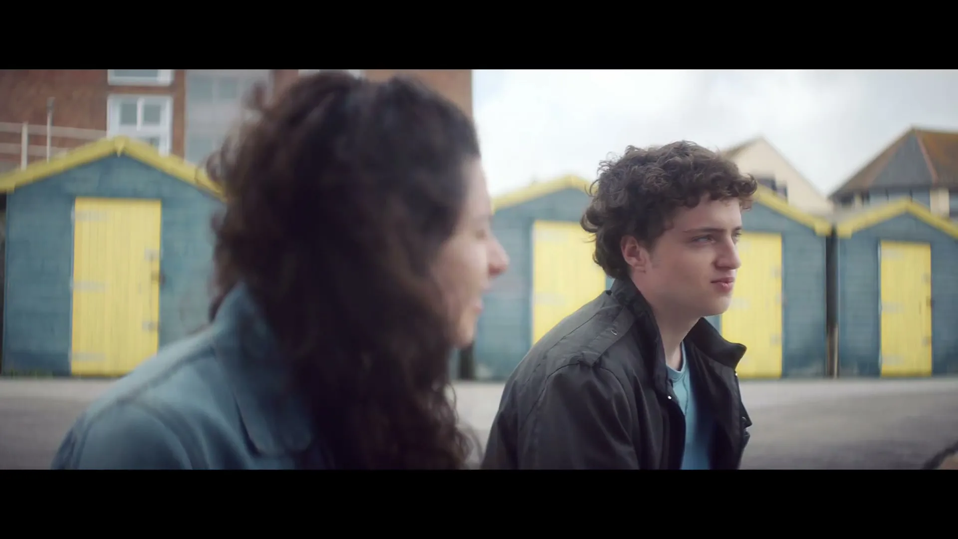 A young man and woman sit on a beach, talking, with colorful beach huts in the background under an overcast sky.