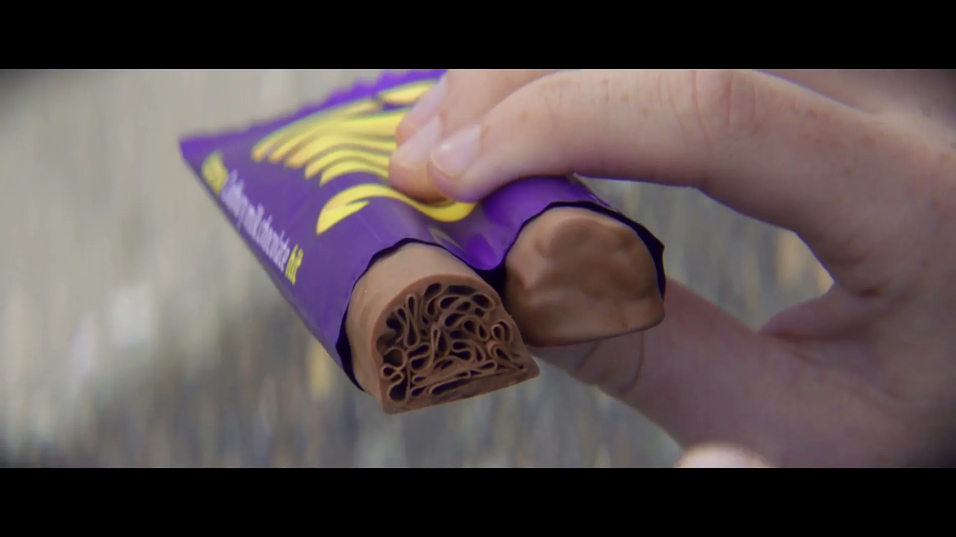 A person's hand holds a Cadbury Twirl chocolate bar, revealing its flaky, aerated interior, against a blurred background.