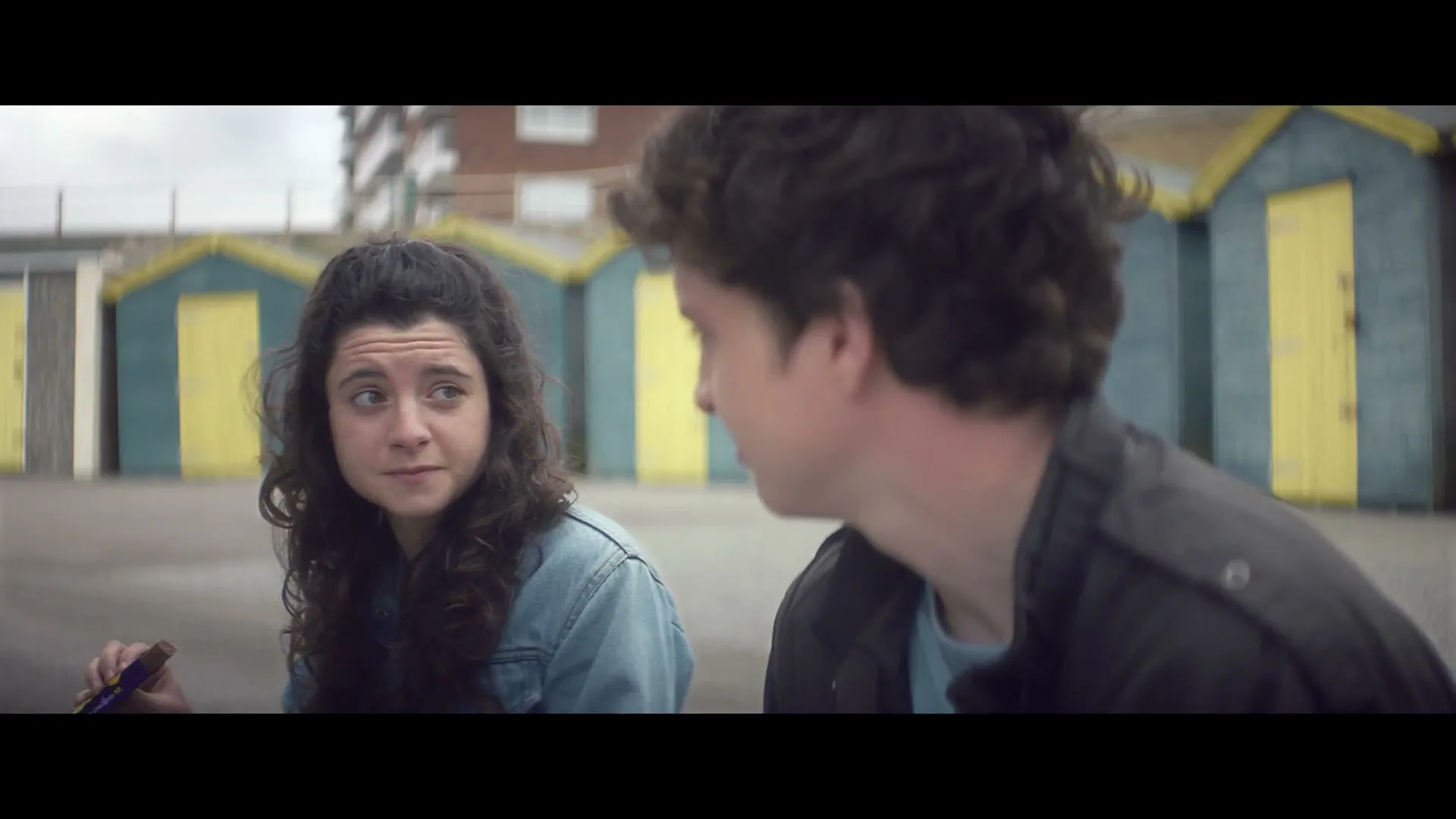 A young woman with curly hair talks to a young man while holding a chocolate bar, with colorful beach huts in the background.